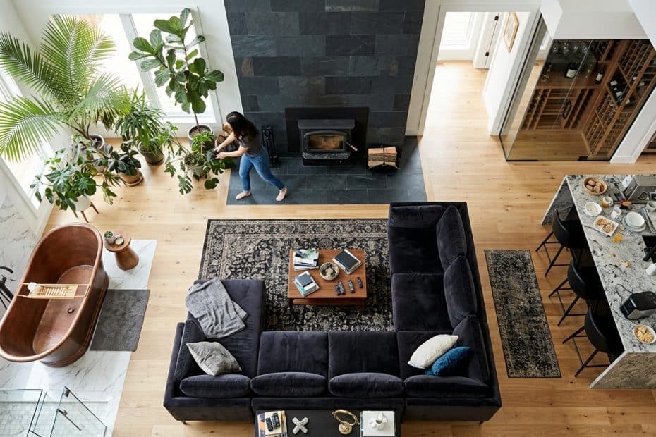 Open-concept living room with dark sectional sofa, wooden coffee table, and a rug; fireplace on a dark stone wall, and a woman tending potted plants by large windows.